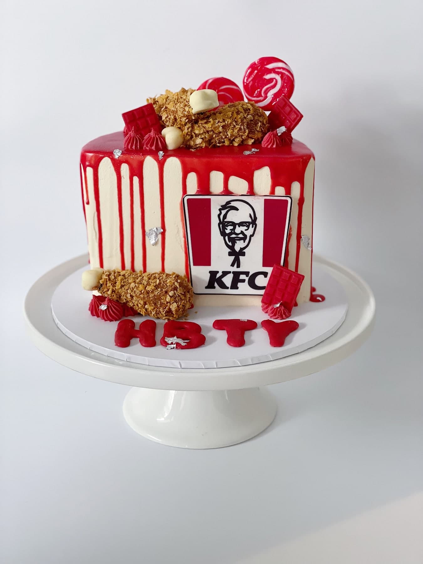 A white cake with a red drip and a KFC logo panel, topped with fried-chicken pieces and red lollies, with a name piped in red at the base.