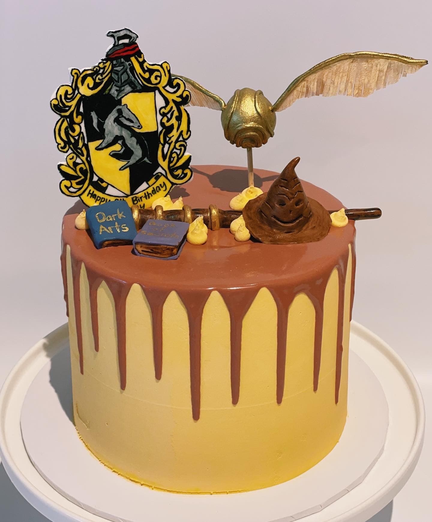 A golden yellow cake with a chocolate drip, topped with a house crest, a winged golden ball, a sorting hat and spell books.