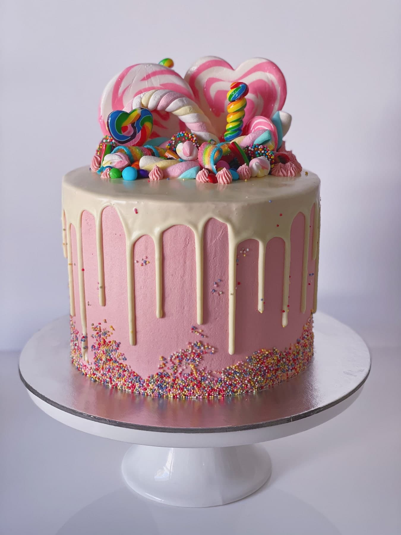 A pink cake with a white drip and rainbow sprinkles, piled high with heart lollipops, marshmallow twists and lollies.