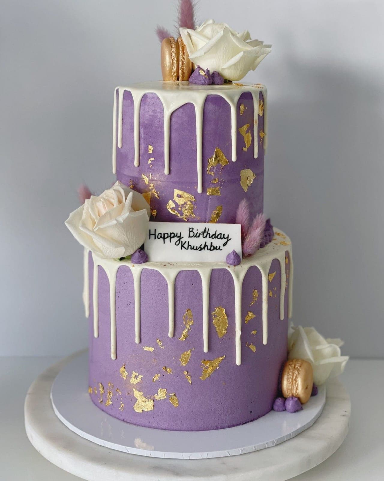 A two-tier lavender cake with a white drip, flakes of gold leaf, white roses, gold macarons and a handwritten birthday plaque.