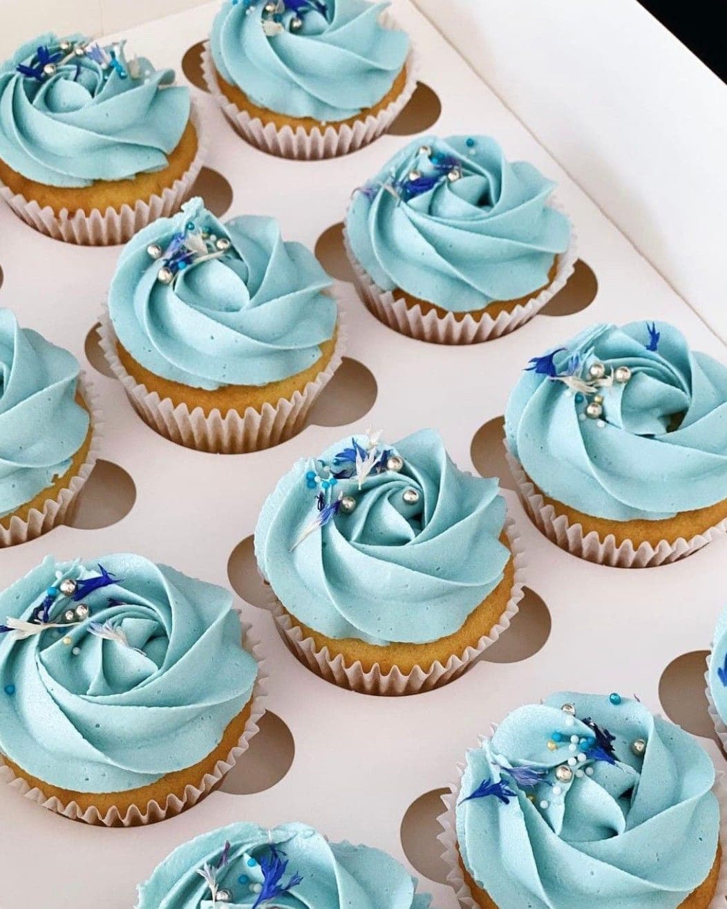 Cupcakes piped with pale blue rosettes, scattered with silver pearls and dried cornflower petals, packed in a box.