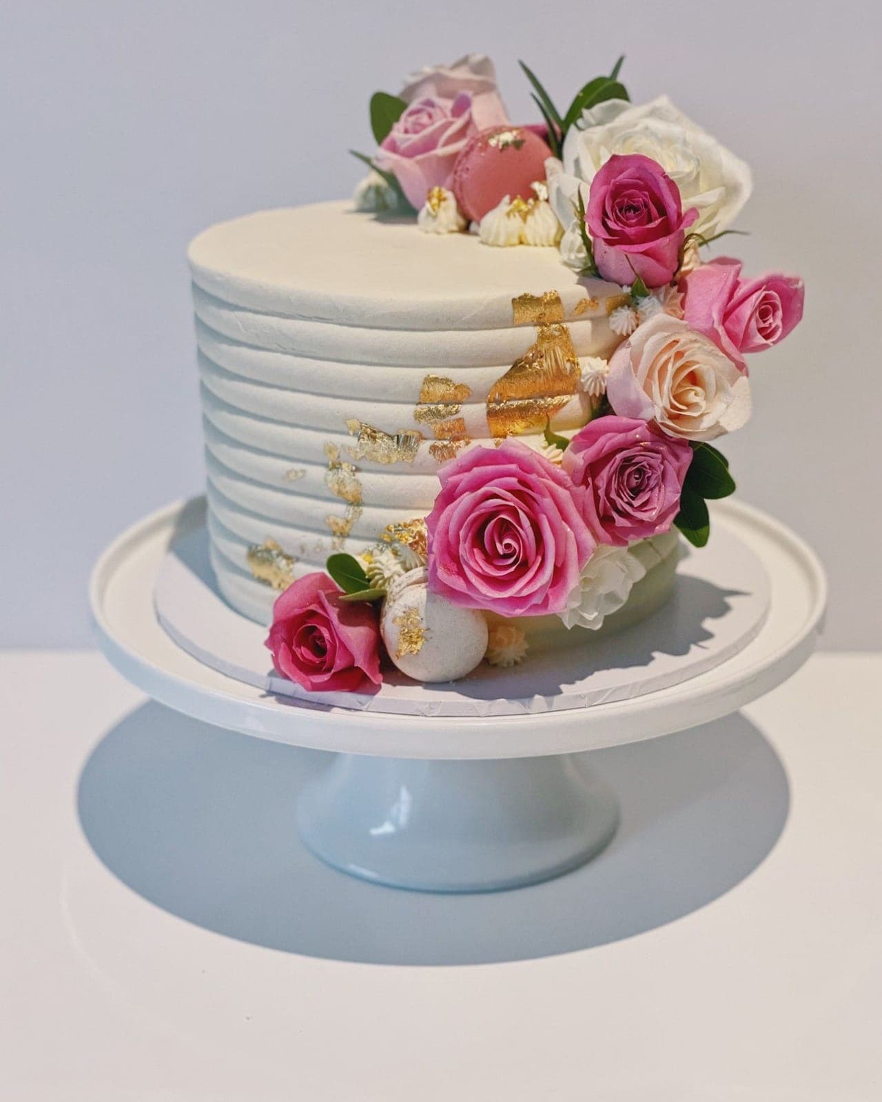 A single-tier ivory cake with ribbed sides and gold leaf, with pink and white roses and macarons cascading down one side.