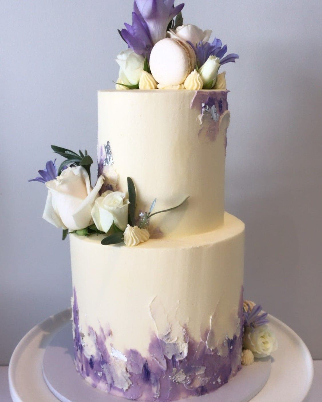 A two-tier ivory cake brushed with lavender and silver leaf around the base, topped with white roses, purple blooms and a white macaron.