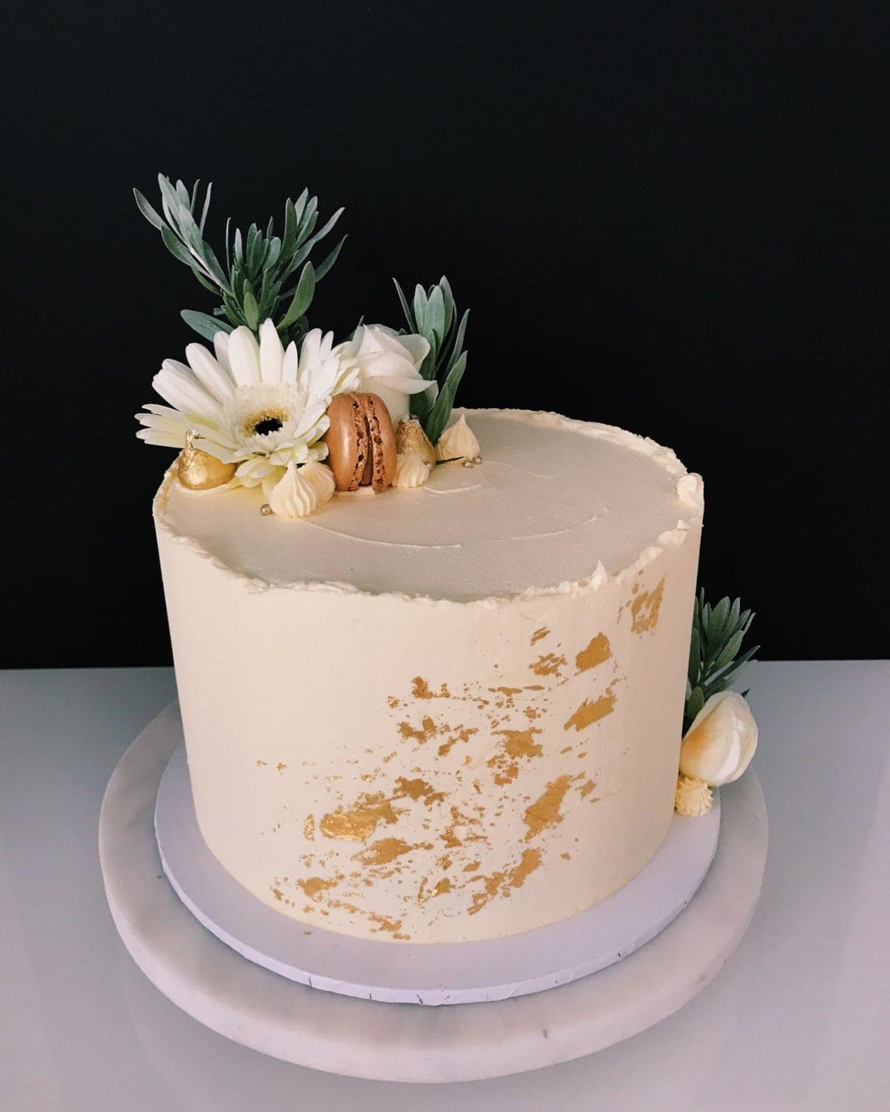 A tall ivory cake with a sweep of gold leaf, topped with a white gerbera, a rose, silvery foliage, a gold macaron and gold-wrapped chocolates.