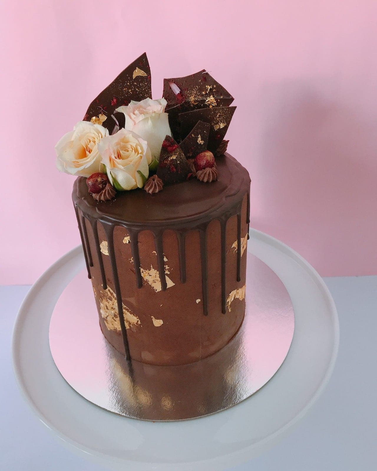 A tall chocolate cake with a dark chocolate drip and gold leaf, topped with chocolate shards and cream roses.