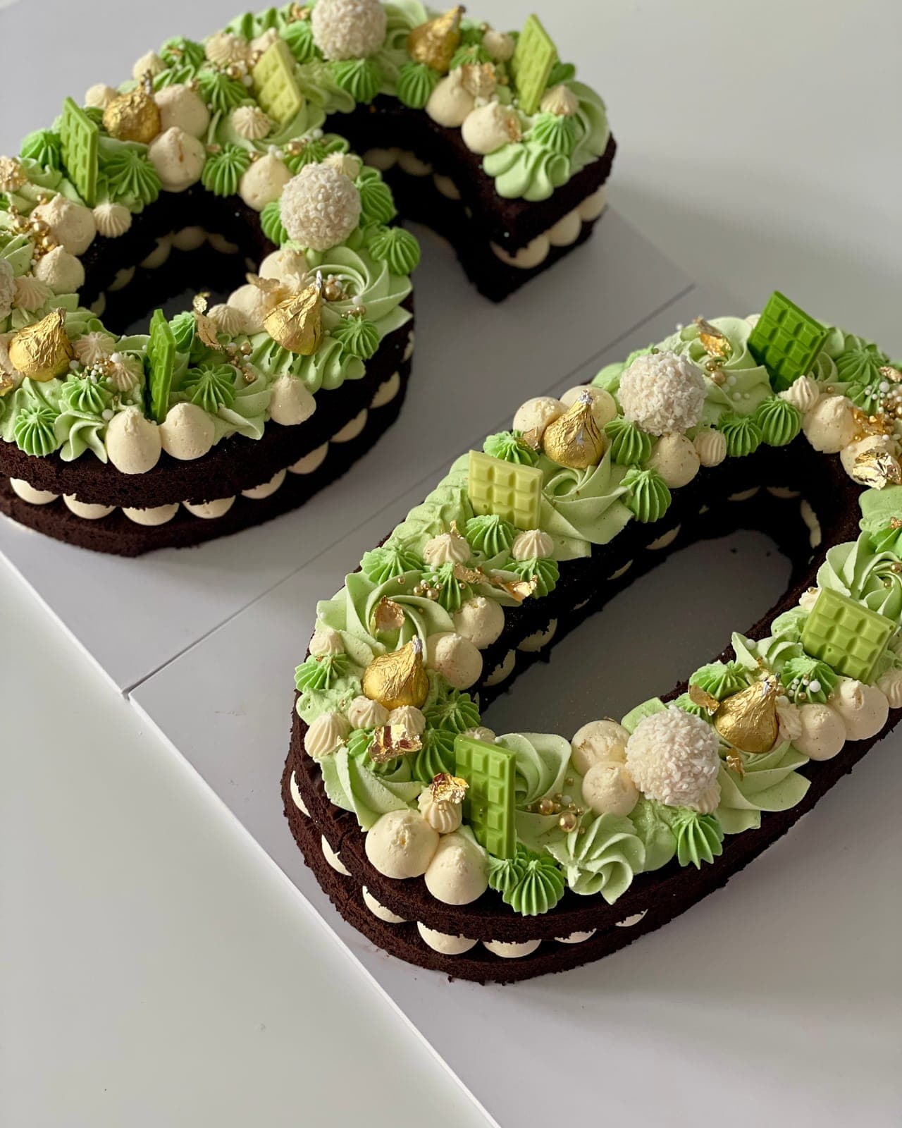 A number cake of chocolate sponge layered with cream, topped with green piped kisses, white truffles, gold chocolates and green chocolate pieces.