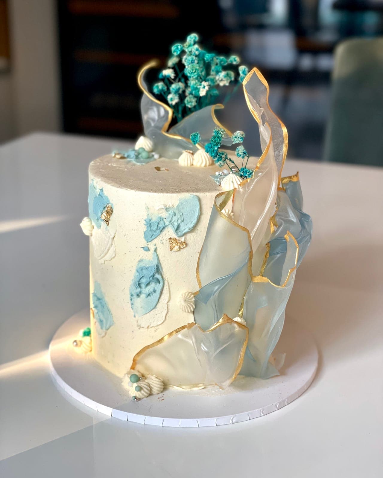 A white cake with blue palette-knife strokes, tall translucent sails edged in gold and a cluster of teal flowers on top.