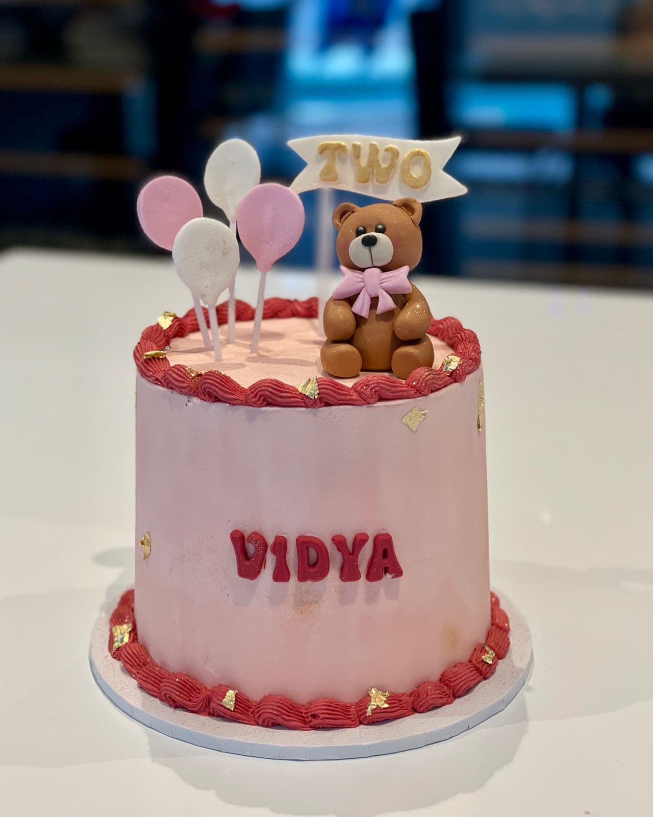 A tall pink cake with a raspberry-red piped border, topped with a modelled teddy bear, pastel balloons and a flag for a second birthday.