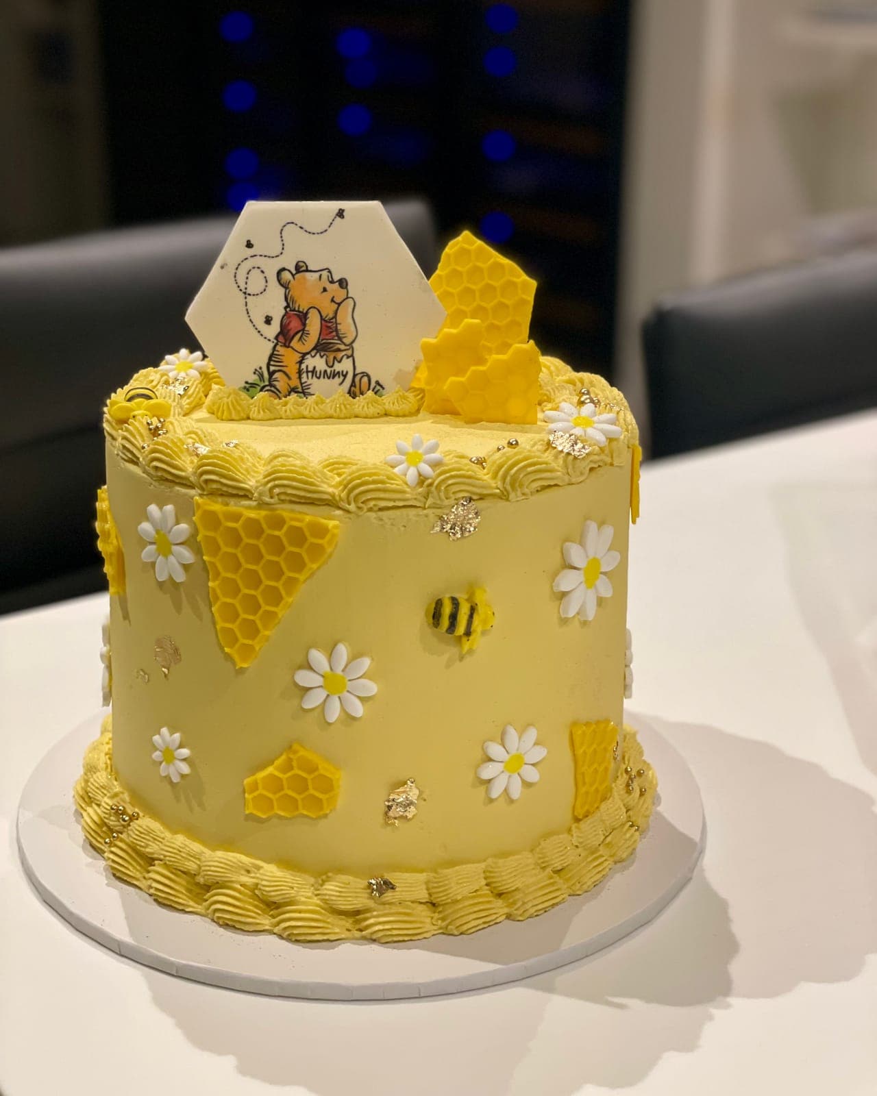 A pale yellow cake with honeycomb panels, sugar daisies and little bees, topped with honeycomb and a storybook bear picture.