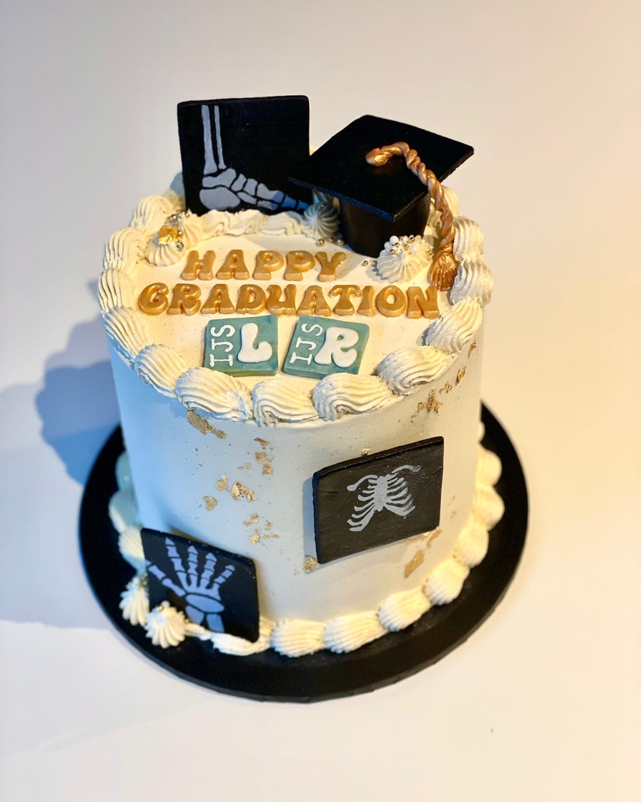 A white cake with gold leaf and black x-ray plaques of a hand, chest and foot, topped with a graduation cap and Happy Graduation in gold letters.